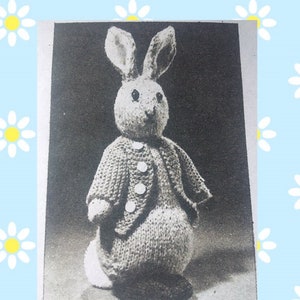 May include: A black and white photo of a knitted bunny rabbit wearing a knitted jacket with buttons. The bunny is sitting on a white surface.