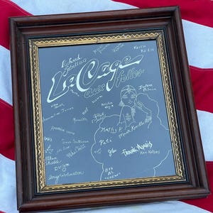 May include: A framed mirror with a gold trim and a blue background. The mirror is covered in white writing, including the title "La Cage aux Folles" and a list of names.