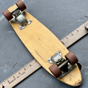 K&ouml;nnte beinhalten: Ein Vintage-Skateboard aus Holz mit Metalltrucks und roten R&auml;dern. Das Skateboard hat eine abgenutzte, verblasste Oberfl&auml;che und liegt auf einer grauen Oberfl&auml;che.