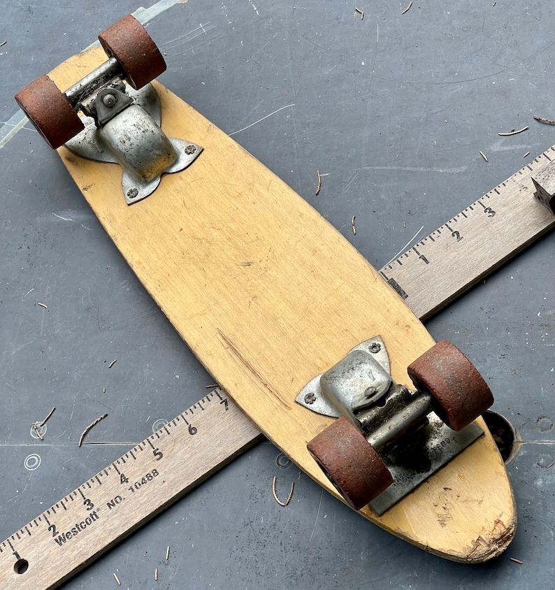 K&ouml;nnte beinhalten: Ein Vintage-Skateboard aus Holz mit Metalltrucks und roten R&auml;dern. Das Skateboard hat eine abgenutzte, verblasste Oberfl&auml;che und liegt auf einer grauen Oberfl&auml;che.