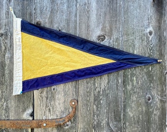 Op de afbeelding: Een gele en blauwe driehoekige vlag met een witte rand en een touw dat aan de bovenkant is bevestigd. De vlag hangt aan een houten muur.