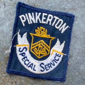 May include: A blue embroidered patch with white stitching that reads "Pinkerton Special Service". The patch features a gold and yellow crest with a stylized flower and leaves.