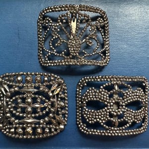 May include: Three antique silver-toned metal buckles with intricate openwork designs. The buckles are square or rectangular, with ornate patterns of beads and embellishments. The background is a solid blue.
