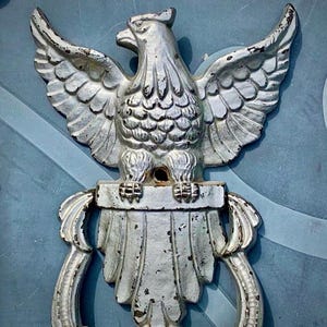 May include: A silver metal eagle door knocker with a detailed design. The eagle is perched on a base with a decorative scroll design.