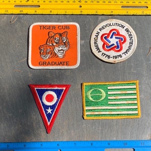 May include: Four embroidered patches. The first patch is orange with a white border and features two tiger cubs with the text "Tiger Cub Graduate". The second patch is white with a red, white, and blue star design and the text "American Revolution Bicentennial 1776-1976". The third patch is a red, white, and blue triangle with a white circle and a star. The fourth patch is green with a white border and features a white circle with a green line through it and white stripes.