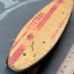 K&ouml;nnte beinhalten: Vintage-Skateboard aus Holz mit roten und wei&szlig;en Streifen und den Worten "Mustang" und "Roller Derby" auf dem Deck.