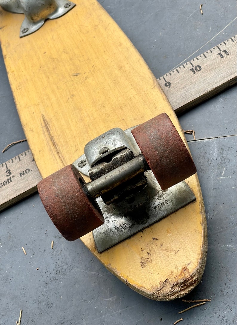 K&ouml;nnte beinhalten: Ein Vintage-Skateboard aus Holz mit zwei roten R&auml;dern und einem Metall-Truck. Das Skateboard hat eine abgenutzte, vergilbte Oberfl&auml;che und einen kleinen Riss an der Unterseite. Der Truck hat eine Metallplatte mit dem Text "PAT. PEND."
