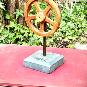 May include: A wooden wheel with five spokes mounted on a black metal rod and a gray square base. The wheel is brown and the base is slate.