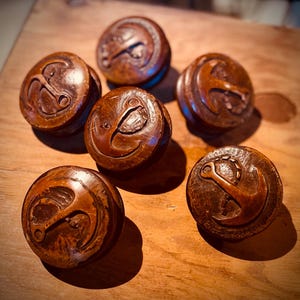 May include: Six round, wooden cabinet knobs with an anchor design carved into the top. The knobs are a warm brown color and appear to be made of a dark wood. The anchor is a lighter shade of brown.