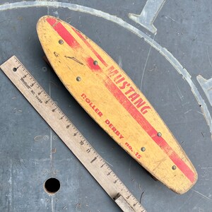 K&ouml;nnte beinhalten: Ein vintage Holz-Roller-Derby-Skateboard mit roten und wei&szlig;en Streifen. Das Skateboard ist mit "Mustang Roller Derby No. 15" beschriftet.