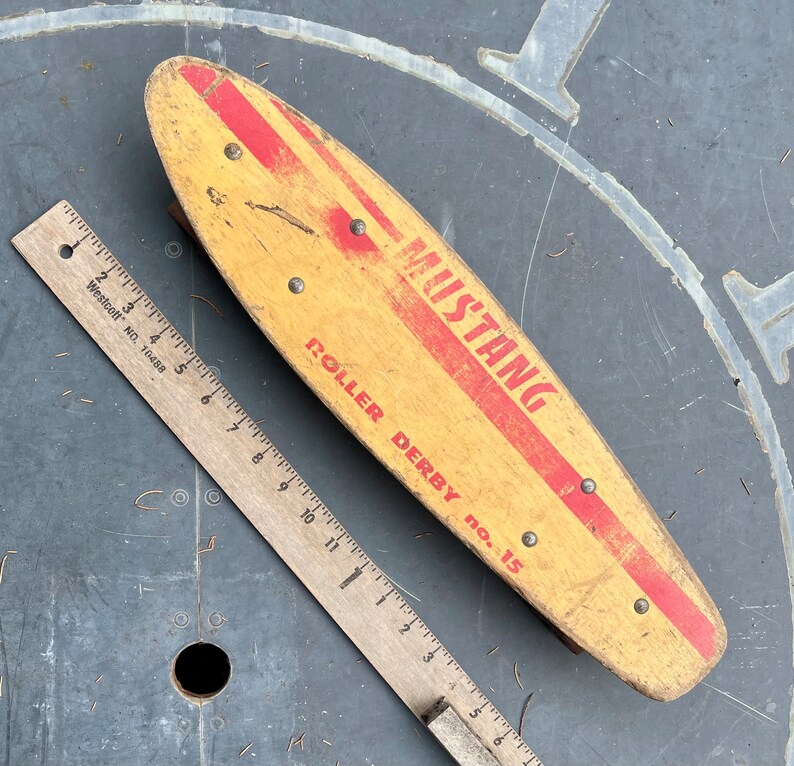 K&ouml;nnte beinhalten: Ein vintage Holz-Roller-Derby-Skateboard mit roten und wei&szlig;en Streifen. Das Skateboard ist mit "Mustang Roller Derby No. 15" beschriftet.
