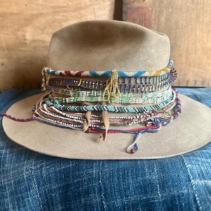 May include: A tan fedora hat adorned with multiple beaded bands in various colors, including red, blue, green, and white. The hat has a wide brim and is set against a backdrop of wooden panels and blue fabric.