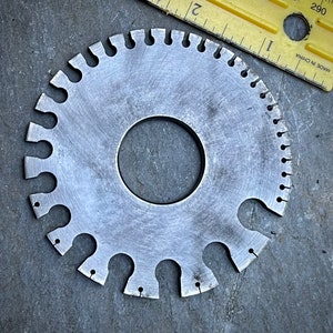 May include: A silver metal gauge with a circular shape and multiple cutouts around the edge. The gauge has a central hole and is designed for measuring the diameter of objects.
