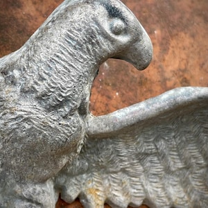May include: Close-up of a weathered, metallic eagle sculpture. The eagle is depicted in profile, with detailed feather textures on its wing. The sculpture has a distressed, aged appearance, with visible signs of wear and tear.