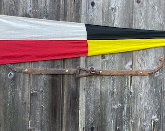 Op de afbeelding: Een witte, rode, zwarte en gele vlag hangt aan een houten deur. De vlag is bevestigd aan een roestige metalen haak.