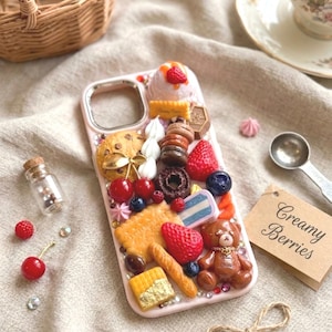 Könnte beinhalten: Eine hellrosa Handyhülle, verziert mit Miniatur-Lebensmitteln wie einem braunen Teddybären, einem Schokoladen-Donut, Macarons, Erdbeeren, Blaubeeren, Kirschen, Keksen und einem Baguette. Die Hülle hat eine metallische Umrandung für die Kamera.