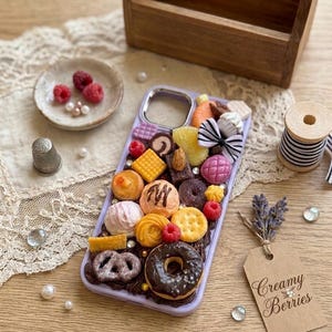 Peut inclure: Une coque de téléphone violet clair décorée de miniatures alimentaires, dont un beignet au chocolat, un bretzel, divers biscuits, des framboises et un nœud rayé noir et blanc. La coque présente une base brune texturée rappelant le glaçage au chocolat.