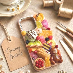 Peut inclure: Une coque de téléphone décorative ornée de breloques miniatures en forme de nourriture, dont une boule de glace verte, un ours en peluche marron, une tarte aux baies, des biscuits et des bonbons gélifiés. Une étiquette porte l'inscription Creamy Berries.
