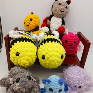 May include: A collection of handmade crochet plush toys, including bees, a ladybug, and other whimsical creatures. The toys are in various colors, such as yellow, black, red, pink, gray, blue, and purple. The toys are displayed on a small wooden shelf.