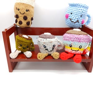 May include: A collection of handmade crochet drink-themed plushies, including a bubble tea, a blue mug, and several coffee cups. The plushies have smiling faces and are displayed on a small wooden shelf. The colors include brown, blue, green, and rainbow.