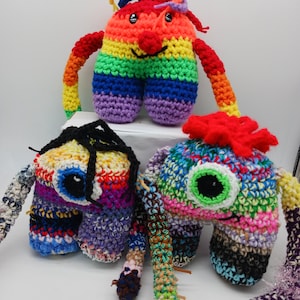 May include: Three handmade crochet plush toys featuring colorful yarn designs. The top toy is rainbow-striped with two eyes. The bottom left toy has black yarn hair and one large eye. The bottom right toy has a red tuft and one large green eye.