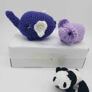 May include: Three crocheted stuffed animals: a purple elephant, a purple pig, and a black and white panda. The animals are sitting on a white box.