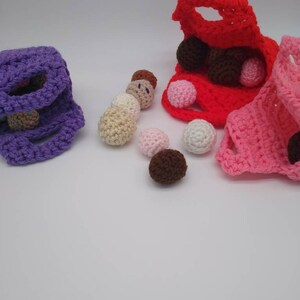 May include: Three crocheted bags in purple, red, and pink, each holding several small, round, crocheted balls in various colors. The balls are scattered around the bags on a white surface. The bags have a simple, open design.