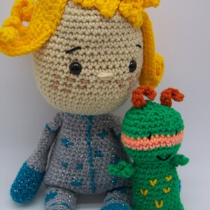 May include: A crocheted doll with yellow hair, a gray and blue outfit, and a green crocheted caterpillar with orange horns.