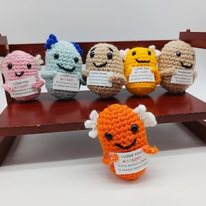 May include: A collection of handmade crochet axolotl plushies in various colors, including pink, blue, brown, yellow, and orange. Each axolotl has a smiling face, small wings, and a tag with the text "I LOVE YOU AXOLOTL". They are displayed on a small wooden shelf.