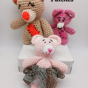 May include: Three handmade crochet teddy bears in shades of tan, pink, and rose. The tan bear has orange accents and a stitched patch. The pink bear has a white nose and a stitched design. The third bear is pink with a gray and brown skirt. The word "Patches" is written in black.