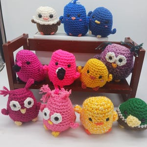 May include: A collection of small, round, crocheted plush figures in various colors including blue, pink, yellow, purple, and green. The figures feature stitched details like eyes and beaks, and some are arranged on a small wooden bench.