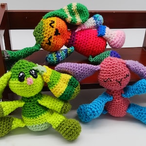 Puede incluir: Cuatro muñecos de conejito de ganchillo coloridos con orejas largas y brazos extendidos. Los conejitos son en tonos de verde, rosa, rojo y naranja, con acentos de azul, morado y amarillo. Están frente a un pequeño banco de madera.