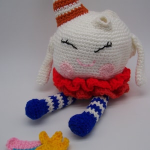 May include: A handmade crochet doll with a white head, pink cheeks, and closed eyes. It wears a red ruffled collar, blue and white striped legs, and a party hat. A small crochet star and rainbow are next to the doll.