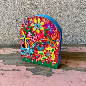May include: A colorful ceramic napkin holder with a red base, blue top, and green trim. The holder is decorated with flowers, leaves, and two animals.