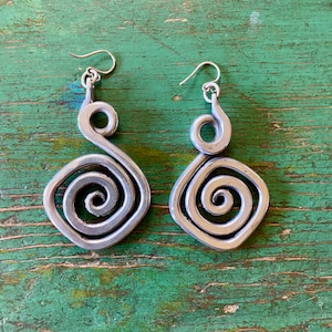 May include: A pair of silver-toned metal earrings. Each earring features a spiral design within a square shape, with a hook for the ear. The earrings are displayed against a green, textured surface.