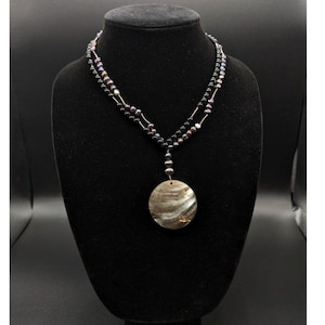 May include: A layered necklace featuring black and iridescent beads, and a large, round shell pendant. The pendant has a swirling pattern of brown, grey, and gold. The necklace is displayed on a black mannequin.