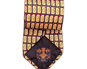 May include: Folded silk tie with a repeating pattern of gold stirrups on a tan background, with red accents and a navy blue border. The tie's underside features a navy blue label with a crest and the words "MORBINO UOMO".