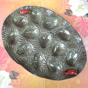 May include: Oval-shaped ceramic serving platter with 12 egg indentations. The platter is glazed in a mottled green-brown color and features a floral pattern. Two small red bird figurines are placed on the rim.
