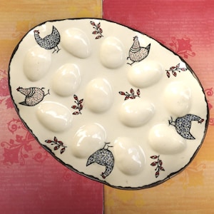 May include: An oval-shaped ceramic egg tray with a cream-colored glaze. The tray features twelve indented egg cups and is decorated with black and white illustrations of chickens and small red and black leaf sprigs scattered across the surface.