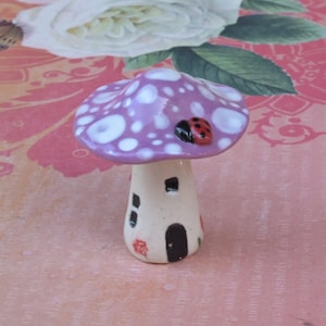 May include: A whimsical ceramic mushroom house with a lavender cap dotted with white spots. A small ladybug rests on the cap. The mushroom stem is off-white with a black door and windows, and red floral accents.