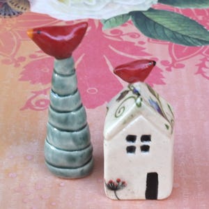 May include: Two ceramic figurines: a tall, spiraled teal form topped with a red bird, and a small white house with a red bird on top. The house has a black door and four square windows. Floral details adorn the roof.