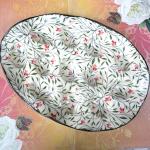 May include: Oval-shaped ceramic serving dish with a cream-colored background. The dish features a delicate pattern of green vines, small red flowers, and a dark rim. The design is reminiscent of a floral garden.