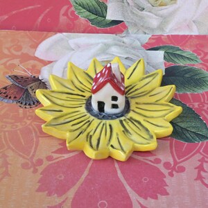 May include: A ceramic miniature house with a red and white polka-dot roof sits in the center of a yellow sunflower-shaped dish. The dish has black lines radiating from the center. The background is a floral pattern with a butterfly.