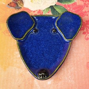 May include: A cobalt blue ceramic dog-shaped dish with a glossy finish. The dish features raised ears, two small black dots for eyes, and a black nose. The edges are outlined in gold. A decorative piece for the home.