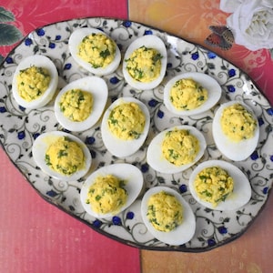 May include: A platter of deviled eggs, a classic appetizer. The eggs are halved, with a yellow filling speckled with green herbs. The white egg halves are arranged on a decorative oval plate with a floral pattern.