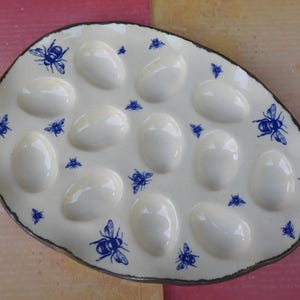 May include: Oval-shaped, off-white ceramic egg serving dish with 12 egg-shaped indentations. The dish is decorated with blue bee illustrations and has a dark brown rim. A decorative serving piece for the kitchen.
