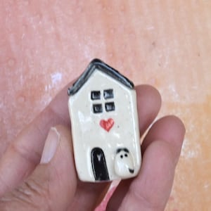 May include: A small, ceramic house figurine with a cream-colored body, black roof, and black door. The house features a red heart and four square windows. A small, white, rounded shape with two black dots is next to the door.