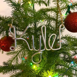 May include: A silver wire ornament spelling "Kyle" hangs on a Christmas tree, next to two red glittery ornaments. The tree is green with colorful lights.