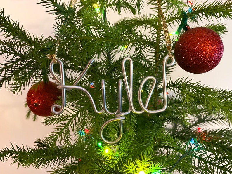 May include: A silver wire ornament spelling "Kyle" hangs on a Christmas tree, next to two red glittery ornaments. The tree is green with colorful lights.