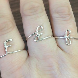 May include: Three silver rings with different designs. The ring on the left finger is a simple band with a letter 'F' design. The ring on the middle finger is a simple band with a letter 'E' design. The ring on the right finger is a simple band with a bow design.